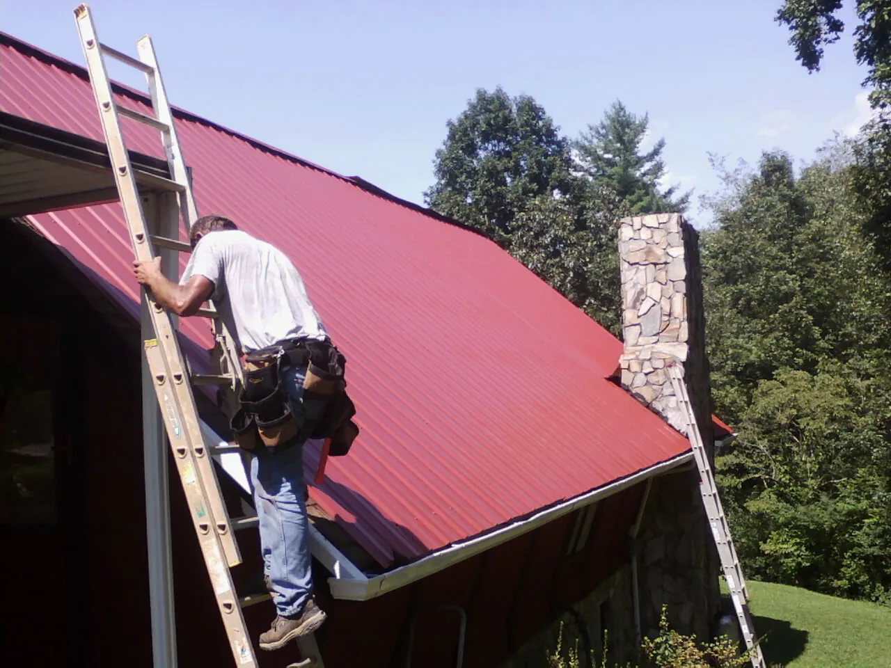 Roofer on a ladder beside a red metal roof and stone chimney — Soffit & Fascia Repair by Roofing Brothers in Rushmore